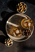 Top view of two spiced cognac cocktails, garnished with cinnamon and star anise, served on a metal tray next to bowls of almonds, peanuts and nuts. The dark, textured background highlights the rich colours of the drink and the nut mixture.