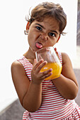 A playful child in a striped top makes a funny face while holding a glass of orange juice. The picture captures the innocence and joy of childhood in an open and lively way.