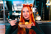 A red-haired woman enjoys Asian street food at a lively market. She is holding chopsticks and a paper tray, surrounded by the bustling atmosphere of the market