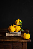 A rustic still life with bright yellow lemons with green leaves, elegantly arranged on a pile of books on a wooden table against a dark background.