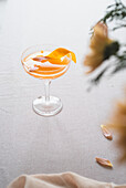 A sophisticated tangerine cocktail adorned with a bright orange peel, served in a classic stemmed glass on a textured white background, accompanied by soft natural light and subtle floral accents.