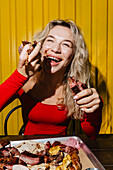 A happy woman in a red top enjoys eating grilled meat with her hands. Sitting in front of a tray of grilled food, she smiles brightly and enjoys the moment in a lively environment.