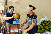 Two Asian women expertly open a coconut to extract the refreshing coconut water and bottle it. The setting is a lively market with a focus on fresh produce and traditional techniques.