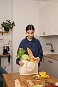 Ein junges Mädchen hält eine Einkaufstüte mit frischem Obst und Gemüse, darunter Salat, Sellerie und Brot, in einer modernen Küche. Die Kulisse betont Gesundheit und Nachhaltigkeit.