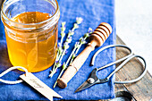 A rustic scene with a jar of honey and a honey spoon on a blue cloth Lavender sprigs and scissors complete the charming arrangement and emphasise the sweetness of nature