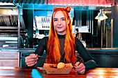 A woman with bright orange hair eats with chopsticks at an Asian market. She sits at a wooden table that shows the culinary culture and the joy of different flavours