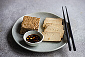 Neatly presented slices of soya tofu, grilled and served on a ceramic plate with a spicy dipping sauce. A pair of chopsticks complements this Asian-inspired dish, offering a taste of traditional cuisine in a modern setting.
