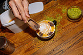 A close-up of a hand finishing off a beautifully prepared speciality coffee with artistic latte art. The wooden table and green powder create a rustic ambience.