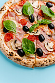 Top view of a freshly baked pizza topped with bacon, mushrooms, mozzarella, black olives, cherry tomatoes and fresh basil on a blue background.