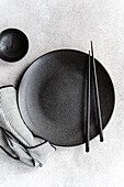 A minimalist black and white placemat with a textured dark plate, a matching bowl, a pair of chopsticks and a striped cloth on a light-coloured background.