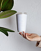 A minimalist white coffee cup floats above a hand in front of a textured white wall A green plant adds a natural accent and emphasises simplicity and elegance