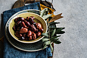 Ceramic bowl filled with Greek grilled olives, accompanied by a strand of fresh olive leaves and a decorative pendant labelled 'Olives'. Set against a rustic background with a vintage napkin, this composition epitomises a delicious Mediterranean snack