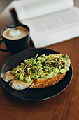 A plate of avocado toast, topped with fresh greens and seeds, is served next to a steaming cup of coffee on a wooden table. An open book lies in the background.