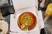 A luxurious cheesecake with crushed pistachios and gold leaf lies elegantly in an open box. The scene takes place in a modern bakery.