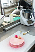 A beautiful pink cake with gold dust lies elegantly on a stainless steel worktop in an Italian bakery, surrounded by baking tools and equipment, ready to be decorated