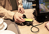 A customer pays with a yellow credit card at a coffee counter showing digital transactions The handheld payment terminal and the beverage cup can be seen on the wooden counter