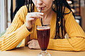 A woman in a yellow jumper sips a refreshing coffee through a straw in a cosy café The focus is on her hands and the drink, creating a warm atmosphere