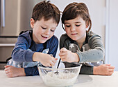 Two young siblings eagerly mix ingredients in a bowl and learn to bake in a kitchen at home. Wearing matching aprons, they demonstrate teamwork and creativity in a fun and educational activity.