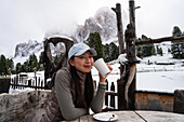 An Asian woman enjoys a warm drink on the Geisleralm in the Italian Dolomites. The rustic surroundings are covered in snow and the picturesque mountains form a breathtaking backdrop.