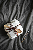 Top view of an elegantly presented bento birthday cake, securely packaged in a white takeaway box decorated with a chocolate brown ribbon. Perfect for gifting, with wooden spoons on a textured grey fabric background.