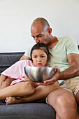 A father and his young daughter sit on a couch at home and share a bowl of popcorn. The scene captures a tender family moment that symbolises love, care and togetherness in a cosy environment.