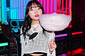 An Asian Gen Z girl having fun in an arcade, holding a large candyfloss The bright neon lights reflect her youthful and trendy style, capturing a joyful moment