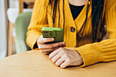 A person in a cosy yellow jumper holds a green smartphone in their hand and engages with the screen The casual setting suggests relaxation or productivity in a café