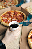 Cropped, unrecognisable male hand holding a white cup of lemonade, with several pizza boxes and a partially visible calzone, all suggesting a casual dining scene.