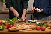 People prepare a fresh sandwich with crispy baguette, cream cheese and various vegetables. Vibrant ingredients and hands in action create a warm, inviting kitchen scene.
