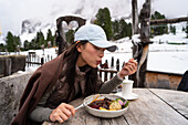 An Asian woman enjoys a hearty meal at the Geisleralm in the snowy Dolomites, Italy Surrounded by rustic wooden structures and trees, she savours the local cuisine in a tranquil setting
