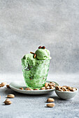 A glass filled with pistachio ice cream, garnished with pistachios and nuts, stands on a plate. It is accompanied by scattered pistachios and a small bowl, all on a textured grey surface.