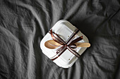 Top view of an elegantly packaged bento birthday cake in a white takeaway box with brown ribbon and wooden cutlery against a background of soft grey fabric.