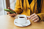A person in a yellow jumper holds a smartphone while enjoying a coffee at a wooden table A white cup, saucer and spoon create a calming atmosphere