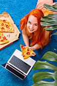 A person enjoys a cosy day indoors, lying on a soft blue carpet with a pizza and a laptop, surrounded by green leaves that create a relaxed and inviting atmosphere