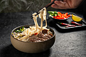 A cut-off, unrecognisable person's hand uses chopsticks to lift steaming, flat rice noodles from a bowl of Vietnamese beef. This aromatic soup contains tender slices of beef, onions and a variety of fresh herbs and offers a flavoursome experience.