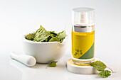 A transparent bottle of birch oil stands next to a white mortar and pestle filled with green leaves against a clean white background that emphasises natural skin care.