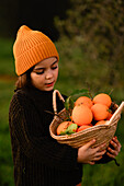 A child in a warm hat and jumper holds a basket full of ripe oranges. The image captures the essence of autumn and natural abundance in a cheerful setting.