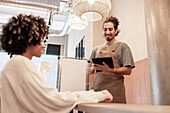 A cheerful coffee house employee with a tablet takes a customer's order The coffee house environment is warm and inviting, with modern lighting and decor - perfect for hospitality themes
