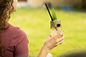 A woman holds a refreshing cocktail with a black straw and garnish in her hand while relaxing outdoors The scene captures a cosy summer moment and embodies relaxation and enjoyment