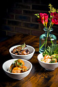 A stylised display of tempura mushrooms in curry sauce and aubergine stew, served with rice in elegant white bowls on a rustic wooden table with floral decorations in the background