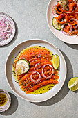 A fresh salmon carpaccio, garnished with slices of lime, cucumber and red onion, sprinkled with spices and sesame seeds. Elegant seafood dish on a light green surface.