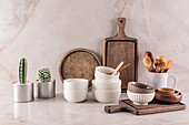 A tidy kitchen worktop with rustic wooden boards, ceramic bowls, wooden utensils and two potted cacti against a marble background.