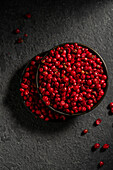 Top view of a dark bowl overflowing with bright red peppercorns against a contrasting, textured dark background. The bright colour of the peppercorns underlines their freshness and spicy flavour.
