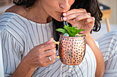 A woman enjoys a refreshing cocktail on a terrace at sunset. She holds a copper cup in her hand and sips through a straw, creating a calm and relaxing atmosphere.