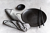 A sophisticated black and white table setting with a round black plate, chopsticks and a striped napkin, representing a minimalist and elegant dining theme.