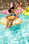 A child relaxes on a donut float in a sunny pool, surrounded by family and friends enjoying inflatable toys. The scene is lively and filled with summer joy.