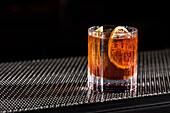 A classic whiskey old fashioned on a metal base. The cocktail contains ice cubes and a slice of dried orange, casting a dramatic shadow and creating an inviting and sophisticated atmosphere.