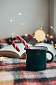 Cosy Christmas ambience with a steaming cup and a candy cane resting on an open book, surrounded by subdued lighting and festive decorations to create a feeling of Christmas warmth and cheerfulness.
