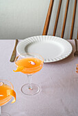A well-appointed dining table with a bright mandarin drink in a sophisticated glass, complemented by a classic white plate and vintage cutlery on a textured tablecloth.