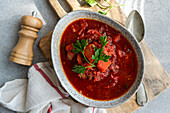 View from above of the traditional Ukrainian beetroot soup Borscht, served in a rustic ceramic bowl, surrounded by fresh herbs and an old pepper mill, taken on a sunny day.
